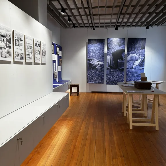 Im Ausstellungsraum eines Museums. An den Raum-Wänden hängen diverse Zeichnungen und Fotografien.