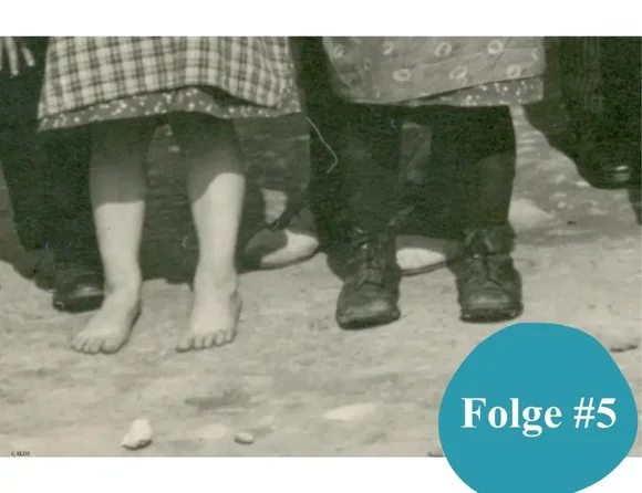 Detail von Kinderfüßen: Das Mädchen ist barfuß, der Junge trägt eine lange Hose und schwarze Schuhe.