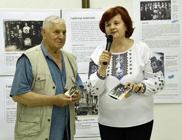 Ein Mann steht neben einer Frau mit einem Mikrofon in der Hand. Beide halten schwarz-weiße Fotografien. Im Hintergrund sind Ausstellungswände zu sehen.