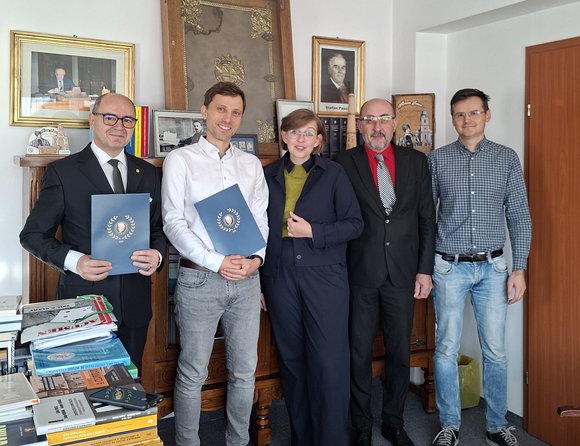 Fünf Personen – vier Männer und eine Frau – stehen in einem Büro nebeneinander und schauen in die Kamera. Zwei Männer halten blaue Mappen in den Händen.