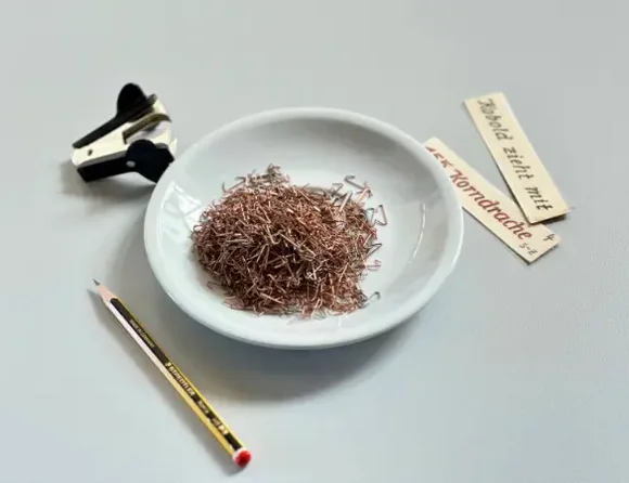 Neben einem Teller mit Tackerklammern liegen ein Tackerklammerentferner, ein Bleistift und zwei kleine Schilder.