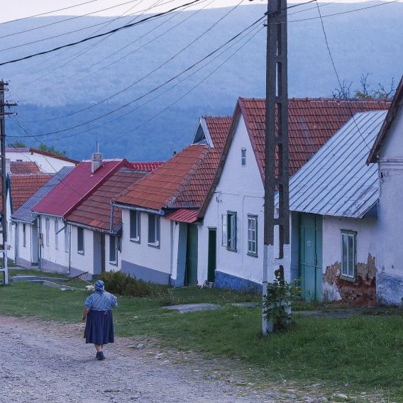 Eine Frau in landwirtschaftlicher Kleidung, trägt einen Rechen über der Schulter und läuft in einem Dorf eine Kiesstraße entlang.