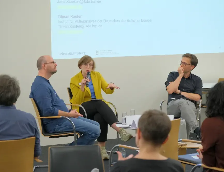 Drei Menschen bei einer Podiumsdiskussion und Zuschauer.