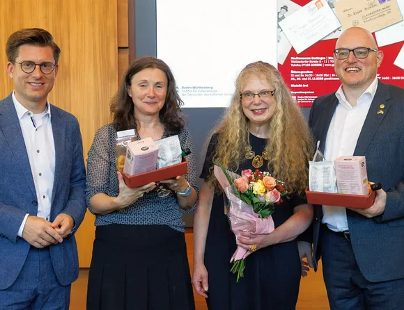 Vier Personen posieren bei einer Ehrung für ein Gruppenfoto. Zwei halten Präsentkörbe, eine Person einen Blumenstrauß.