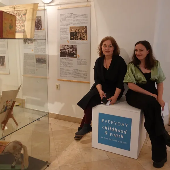 Ein Museumsraum mit Exponaten in Glasvitrinen. Auf einem Sockel sitzen zwei Frauen.