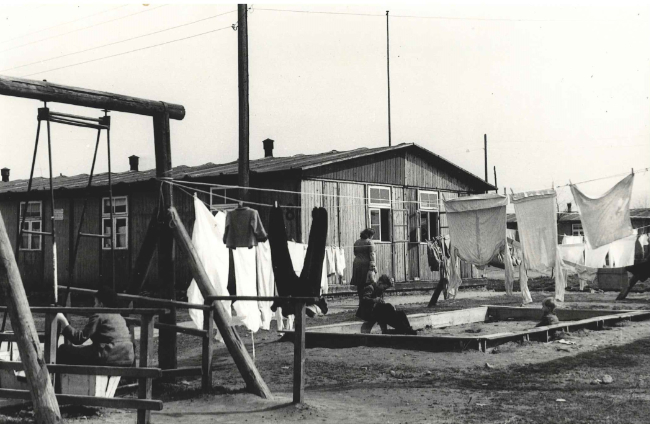 Vor einer Holzbaracke hängt Wäsche auf gespannten Wäscheleinen. Daneben stehen eine Schaukel und ein Sandkasten mit spielenden Kindern.