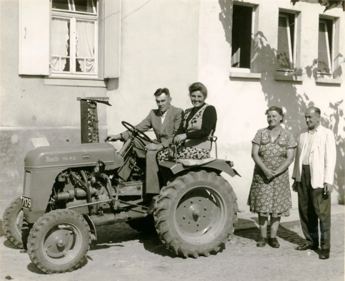 Ein Mann und eine Frau sitzen auf einem Traktor vor einem Haus, daneben stehen ein weiterer Mann und eine Frau.