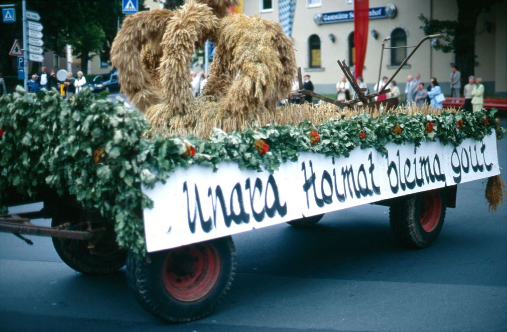 Ein mit grünen Pflanzen geschmückter Festwagen. Darauf eine Krone aus Stroh. An der Seite ein Transparent mit einem Schriftzug im Dialekt.
