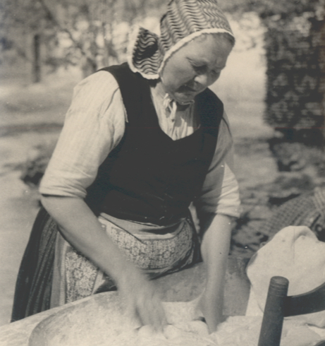 Eine ältere Frau in Arbeitstracht knetet Teig mit den Händen in einem Holztrog.