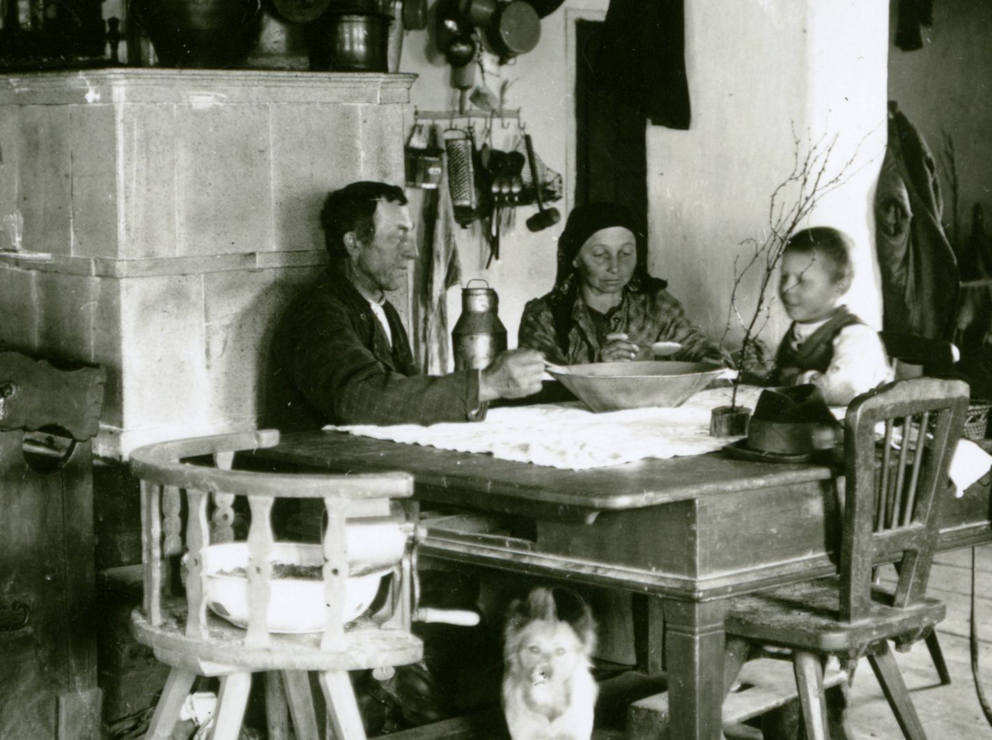 Schwarzweißfotografie eines Raumes. Eine dreiköpfige Familie sitzt in einer Stube um einen Tisch. Unter dem Tisch liegt ein Hund, im Hintergrund steht ein Ofen.