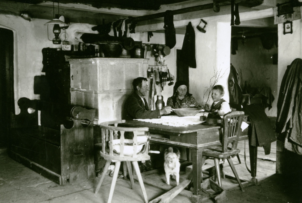 Schwarzweißfotografie eines Raumes. Eine dreiköpfige Familie sitzt in einer Stube um einen Tisch. Unter dem Tisch liegt ein Hund, im Hintergrund steht ein Ofen.