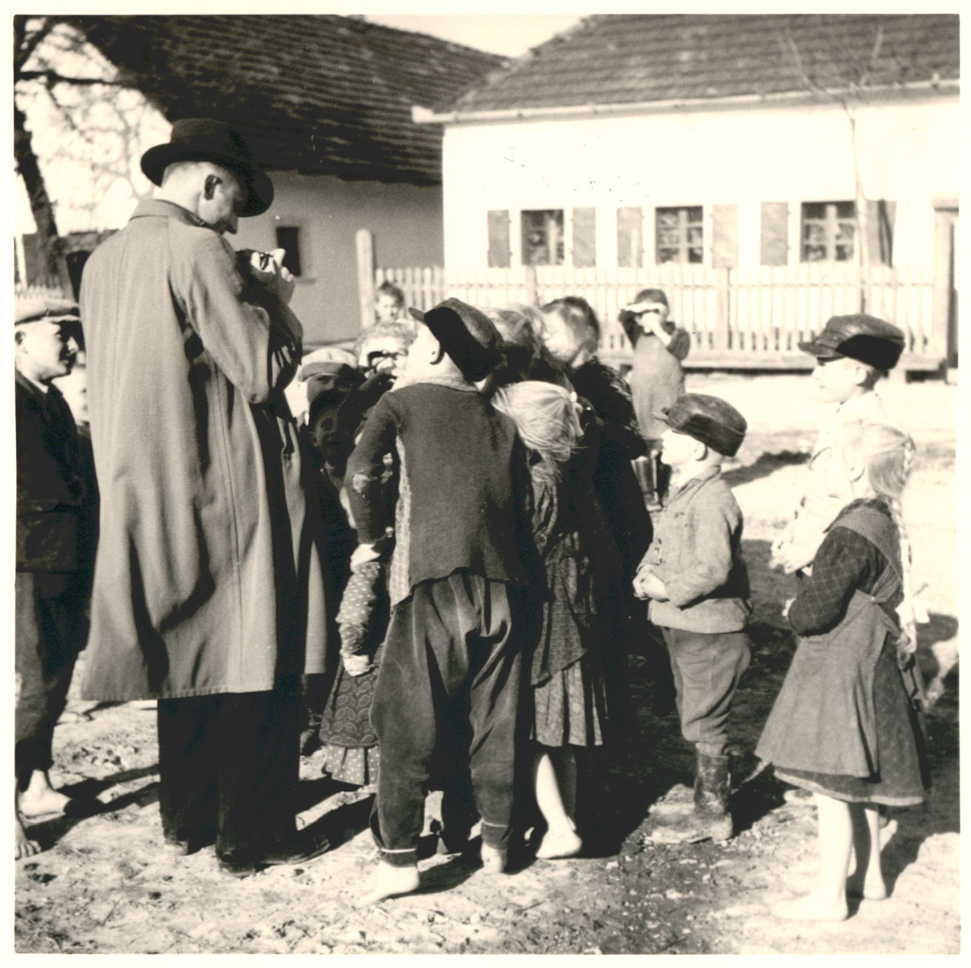 Ein Mann mit Hut und Mantel (vermutlich Hartmann) zeigt seine Kamera einer Gruppe von Kindern, die sich neugierig um ihn scharren