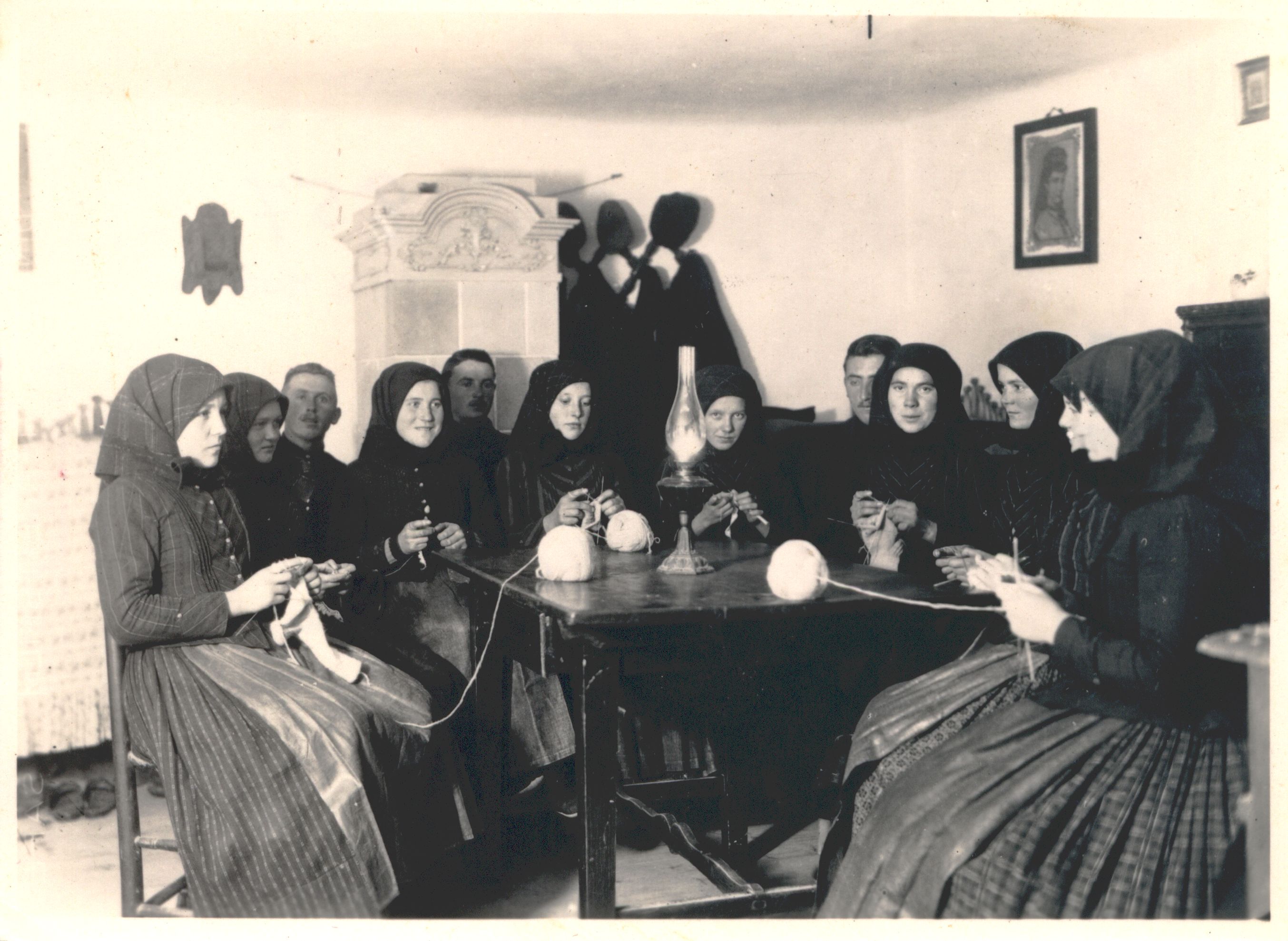 Acht Frauen sitzen um einem Tisch herum und stricken, hinter ihnen sitzen ebenfalls drei Männer. Hinter dem Tisch an der Wand hängt neben einem Ofen eine Garderobe mit Kleidern und Hüten, an der anderen Wand hängt das Porträt einer Frau. 