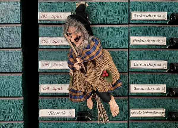 Eine Hexenpuppe hängt vor einem Regal mit grünen Schubern.