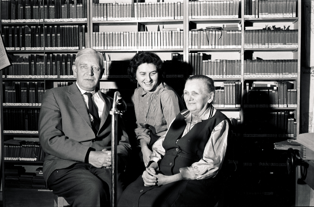 Ein Mann mit zwei Frauen vor einem Mikrofon; im Hintergrund Bücherregale.