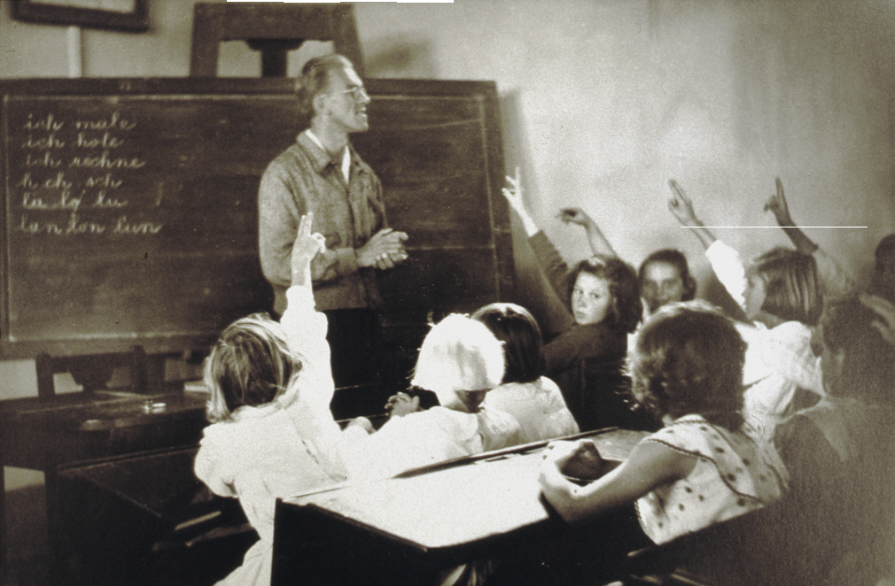 Blick in einer Schulklasse. Ein Lehrer steht vor einer Tafel. In den Schulbänken sitzen Kinder.