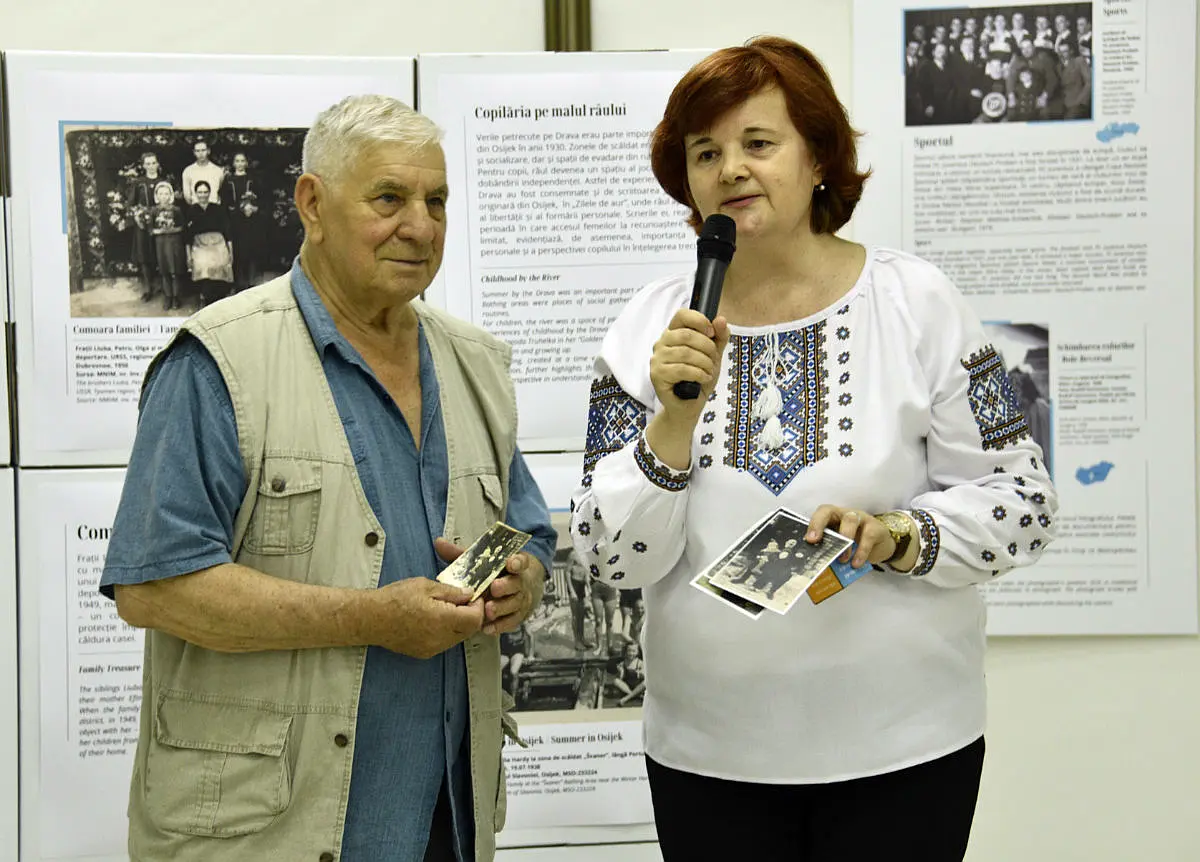 Ein Mann steht neben einer Frau mit einem Mikrofon in der Hand. Beide halten schwarz-weiße Fotografien. Im Hintergrund sind Ausstellungswände zu sehen.