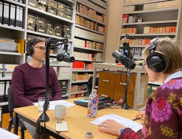 Mann und Frau sitzen sich gegenüber an einem Tisch und sprechen mit Kopfhörern in Mikrofone.