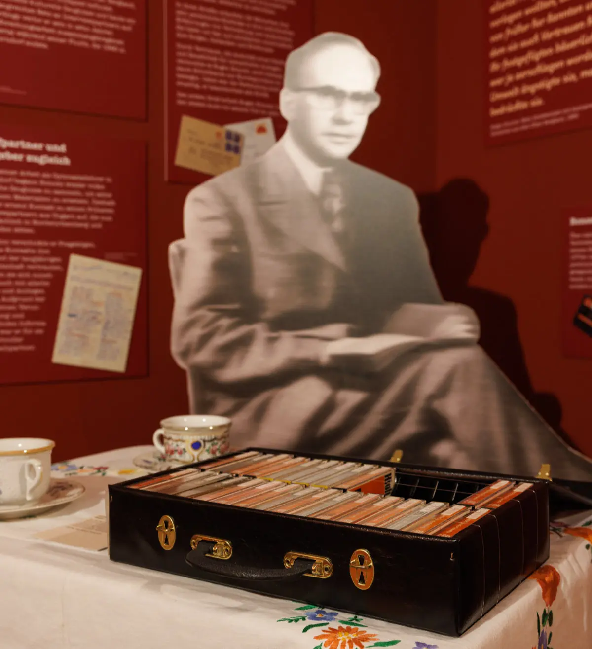 Blick in einen Ausstellungsraum. Zu sehen ist ein großes Schwarz-Weiß-Foto eines sitzenden Mannes. Auf dem Tisch vor ihm stehen ein offener Koffer mit Kassetten sowie zwei Porzellantassen.