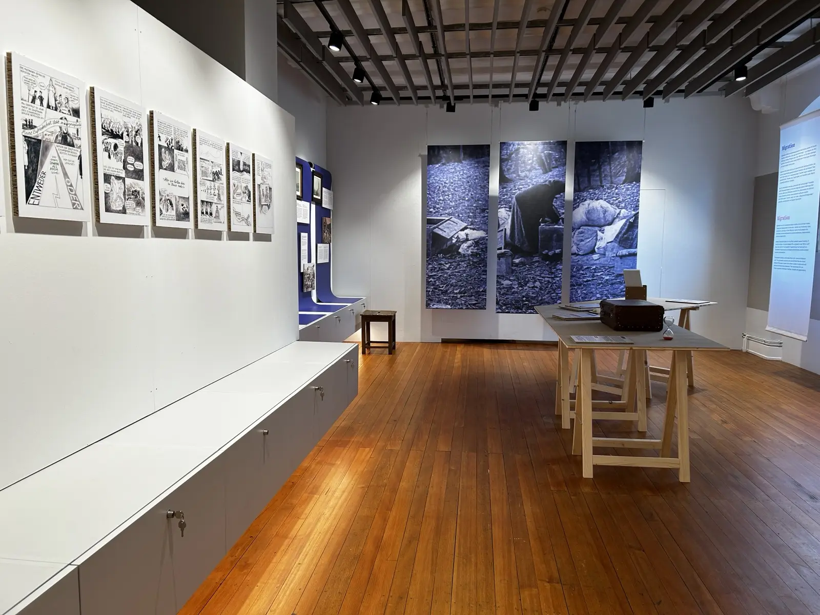 Im Ausstellungsraum eines Museums. An den Raum-Wänden hängen diverse Zeichnungen und Fotografien.