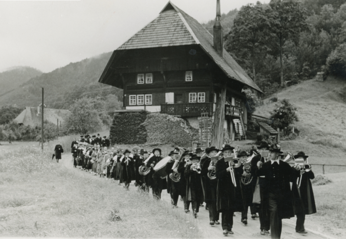 Ein Umzug mit einer Blaskapelle auf einem Weg vor einem Schwarzwaldhaus