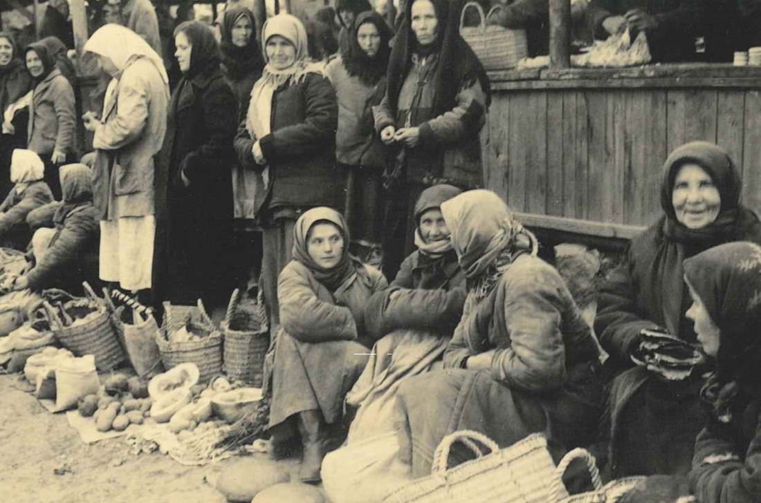 Auf der Straße vor einem Haus sitzen und stehen Frauen; auf dem Boden stehen geflochtene Körbe und Gemüse