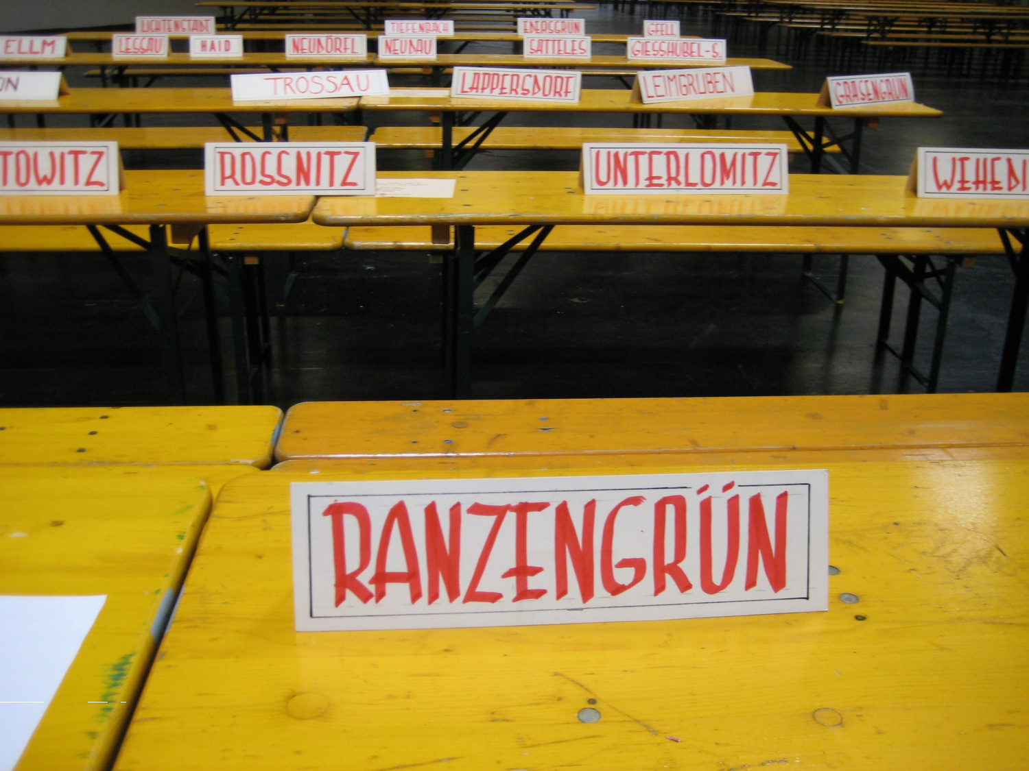 Eine Halle mit aufgereihten Holztischen. An den Tischen Schilder mit Ortsnamen.