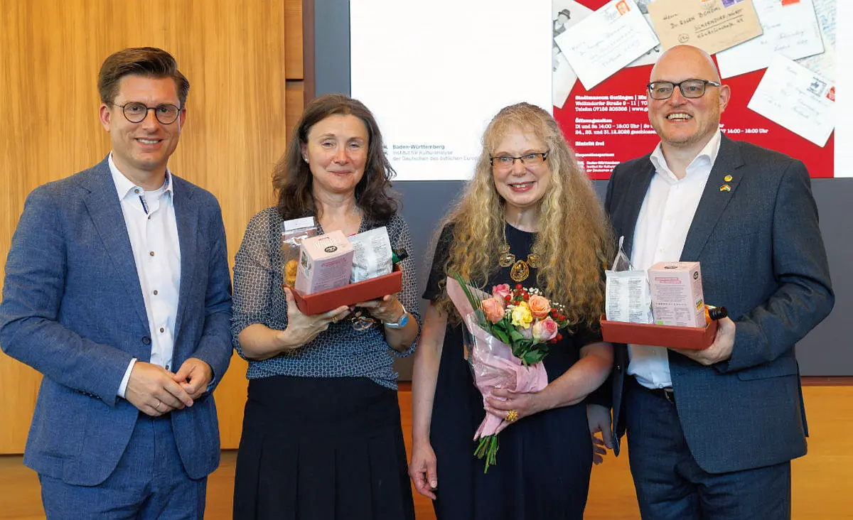 Vier Personen posieren bei einer Ehrung für ein Gruppenfoto. Zwei halten Präsentkörbe, eine Person einen Blumenstrauß.