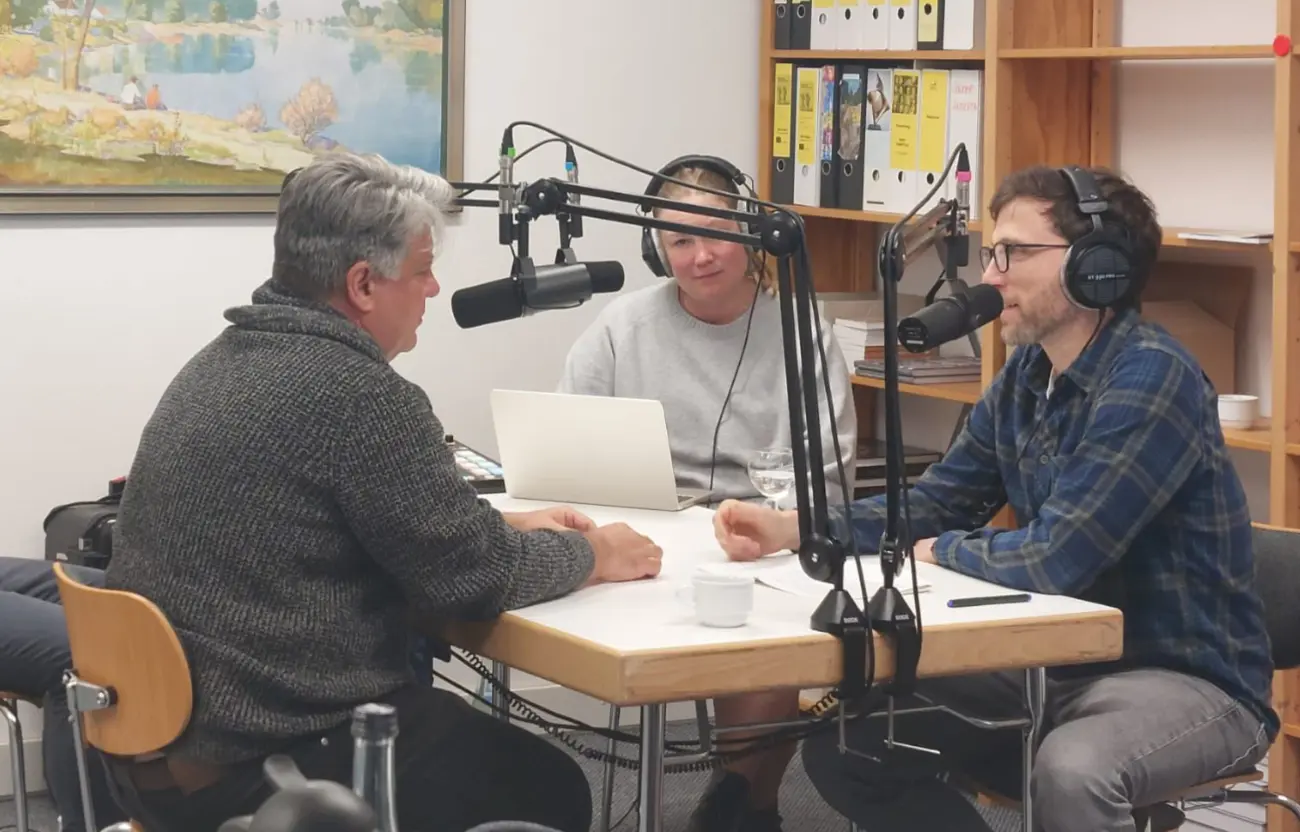 Zwei Männer sitzen an einem Tisch und sprechen in Mikrofone. Eine Frau mit Kopfhörer sitzt daneben..