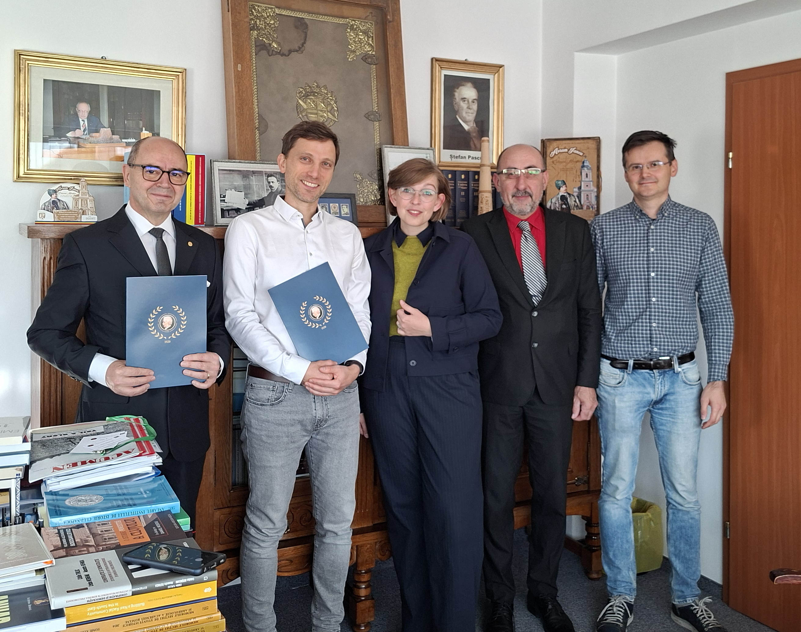 Fünf Personen – vier Männer und eine Frau – stehen in einem Büro nebeneinander und schauen in die Kamera. Zwei Männer halten blaue Mappen in den Händen.