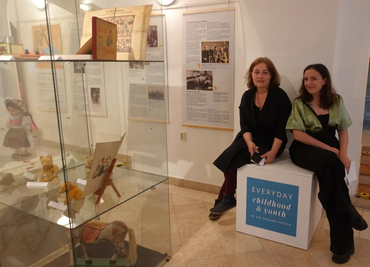 Ein Museumsraum mit Exponaten in Glasvitrinen. Auf einem Sockel sitzen zwei Frauen.