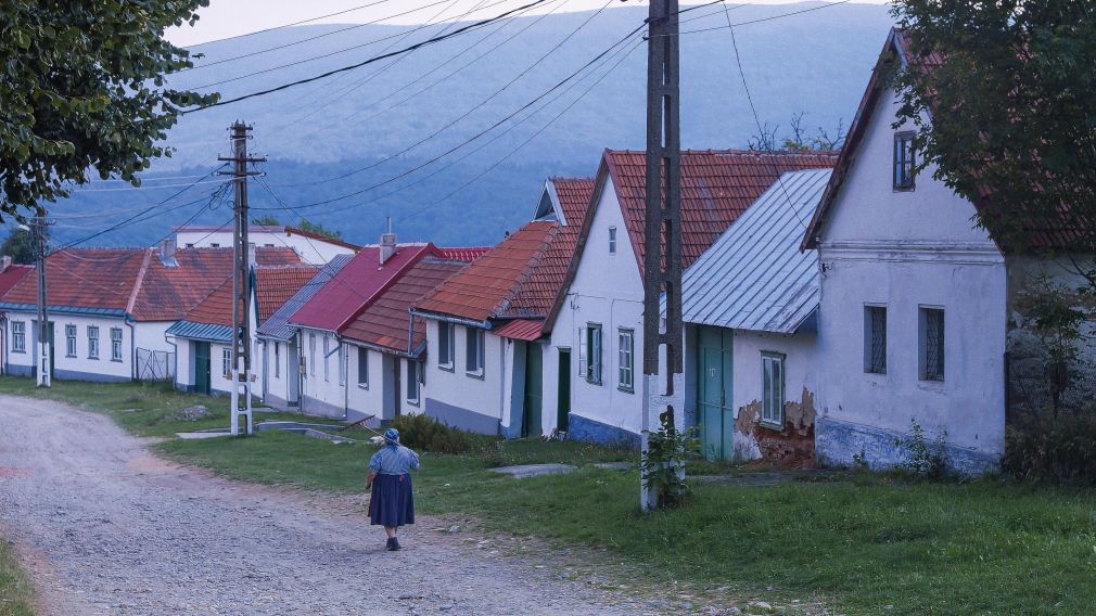 Eine Frau in landwirtschaftlicher Kleidung, trägt einen Rechen über der Schulter und läuft in einem Dorf eine Kiesstraße entlang.