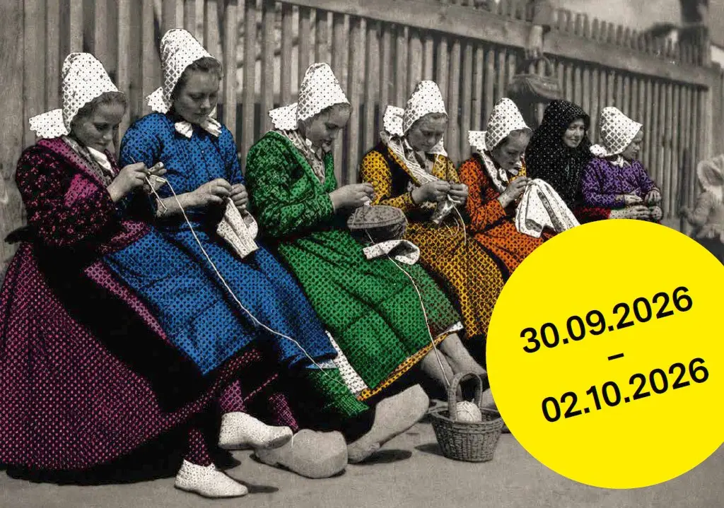 In einer Reihe im Freien sitzende Frauen in Trachten stricken. Die Schwarz-Weiß-Fotografie ist nachkoloriert.